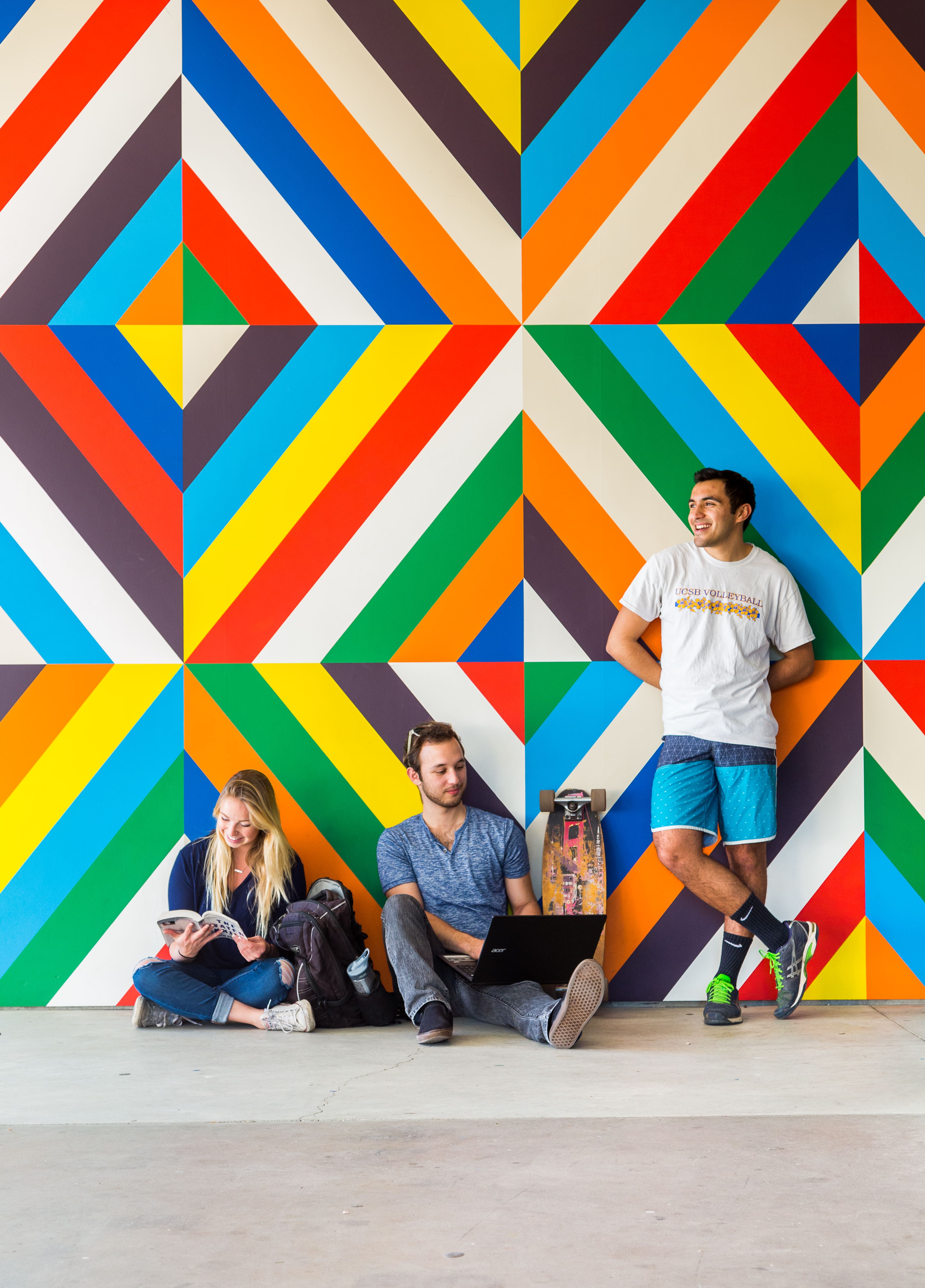 Three Students sitting and standing in front of a geometric, colorful wall.