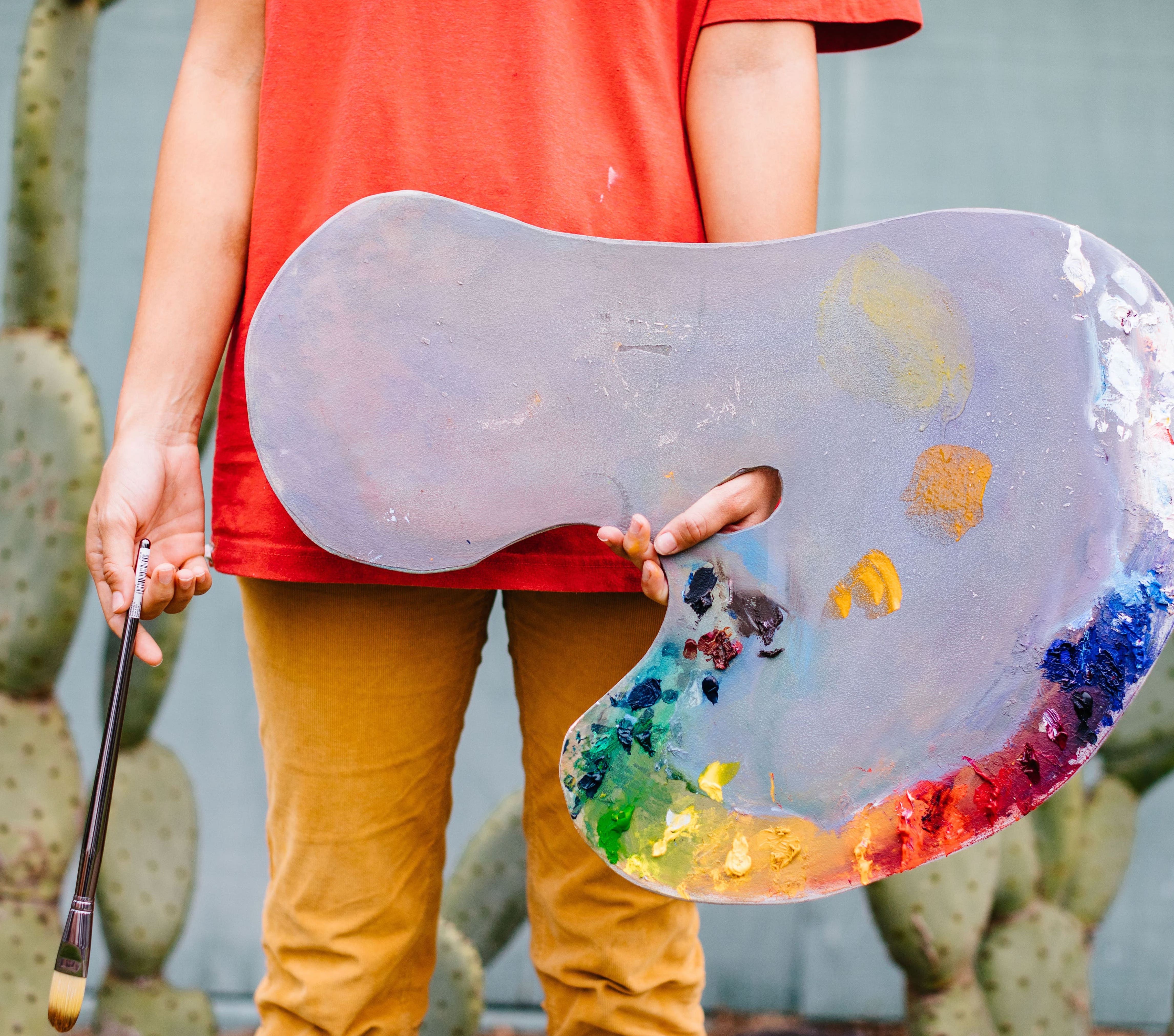 Person wear a red shirt and yellow pants while holding a paint palette in their left hand and hold a paint brush in their right hand. Cacti are in the background