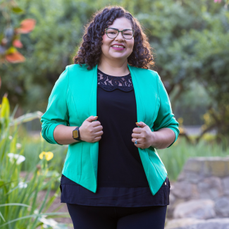 Martha standing in front of greenery, with a green blazer, a watch, glasses, and a big smile. 